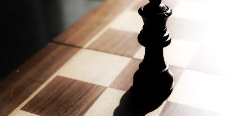 Chess black stands on chess board against the light source with shadow reflected on chess board.