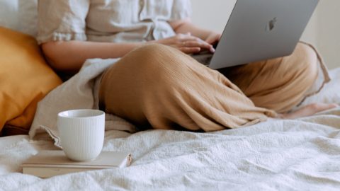 One person uses their laptop on their bed, with a mug on top of a book.