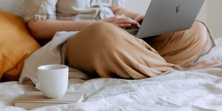 One person uses their laptop on their bed, with a mug on top of a book.