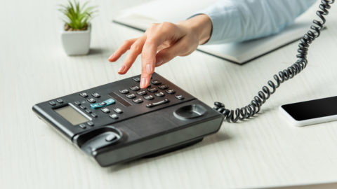 Cropped view of businesswoman dialing number on landline telephone on table with notebook and plant