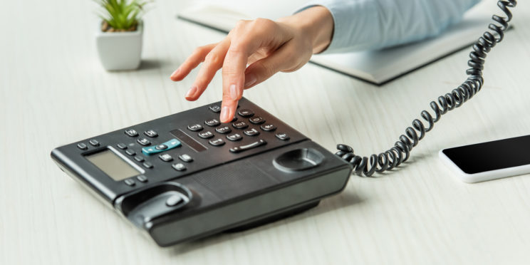 Cropped view of businesswoman dialing number on landline telephone on table with notebook and plant