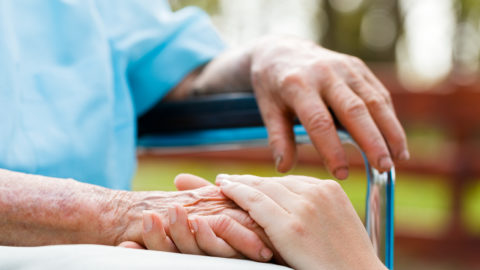 A person is putting their hand on an elder's hand who's sitting in a wheelchairs