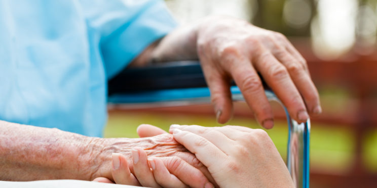 A person is putting their hand on an elder's hand who's sitting in a wheelchairs