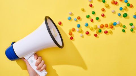 Cropped view of woman holding loudspeaker with colorful candies on yellow background