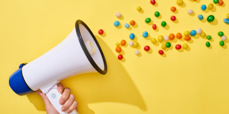 Cropped view of woman holding loudspeaker with colorful candies on yellow background